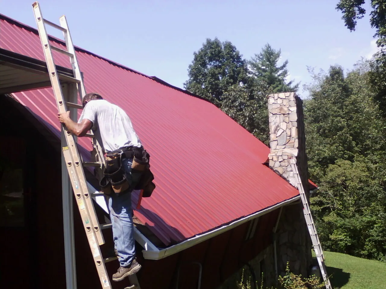 Roofer on a ladder beside a red metal roof and stone chimney — Soffit & Fascia Repair by Roofing Brothers in Fleischmanns